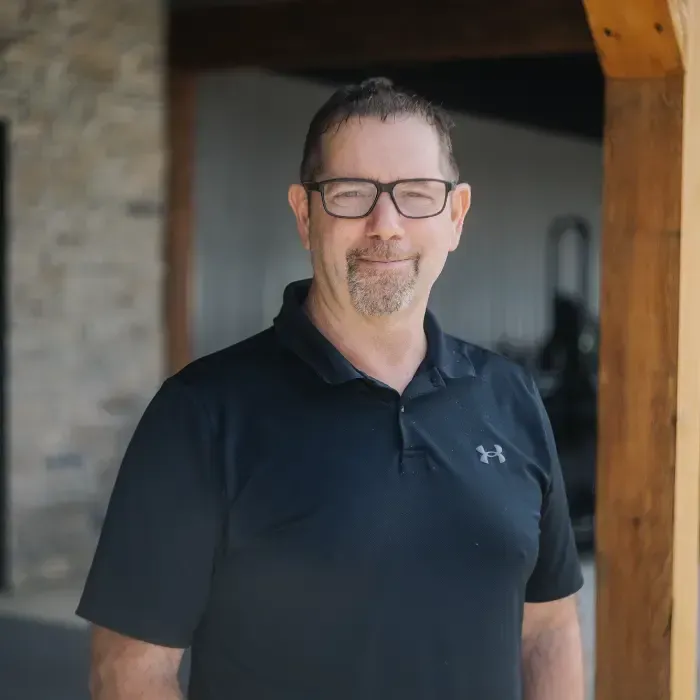 Man in black polo and glasses standing outdoors under a wooden beam