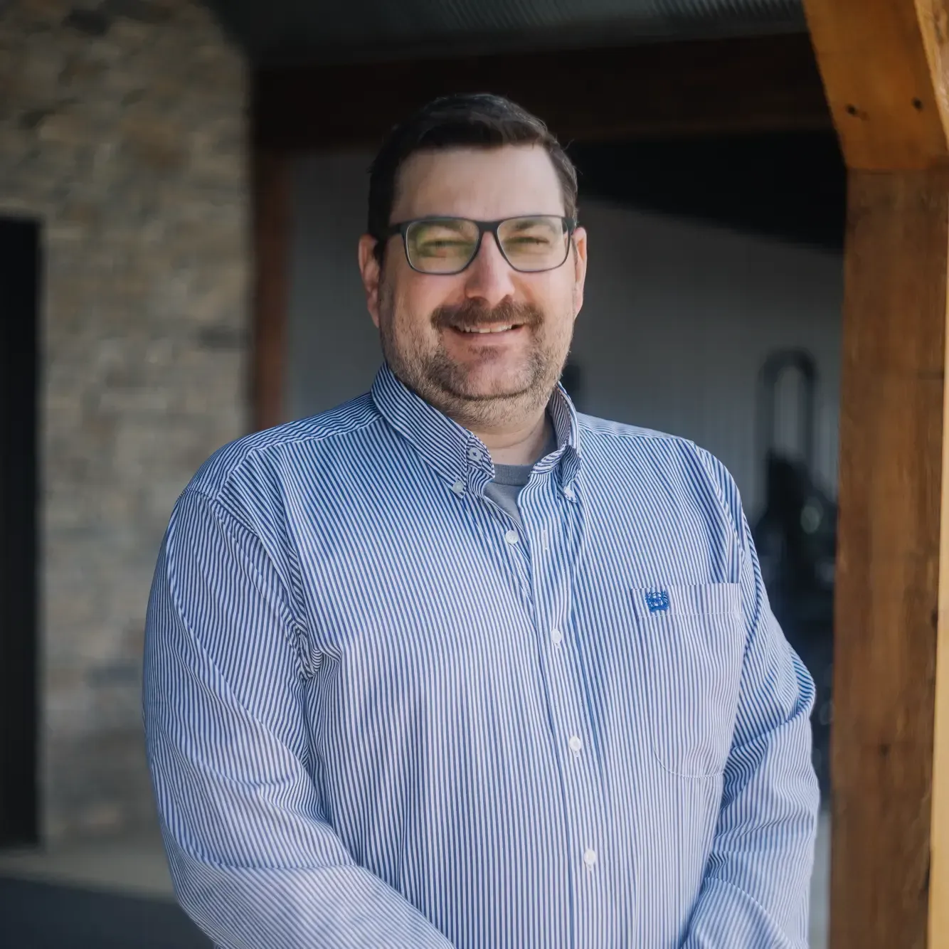 Man wearing glasses and a blue striped shirt standing and smiling indoors by a wooden post
