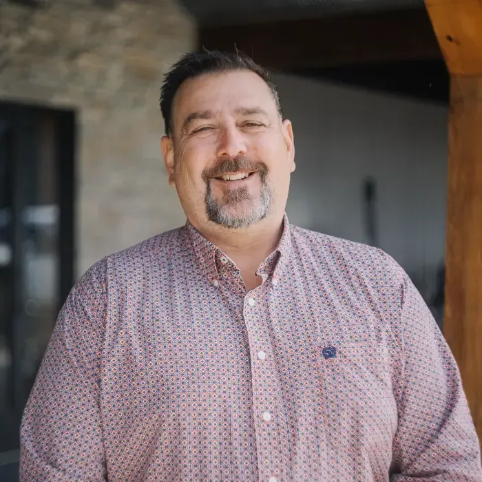 Smiling man in a patterned shirt standing outdoors in front of a stone wall
