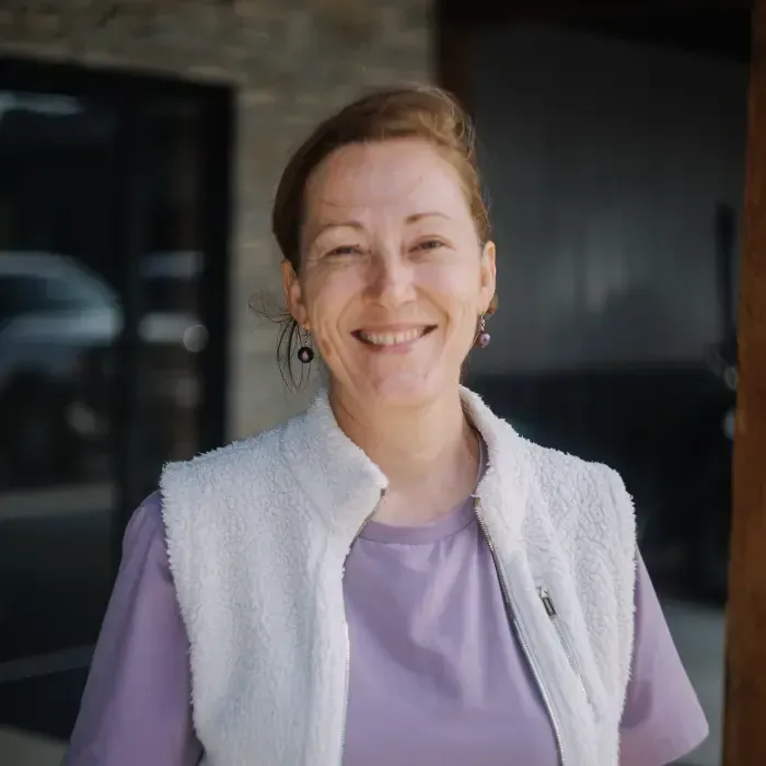 Smiling person in a lavender shirt and white vest standing outdoors in front of a building.