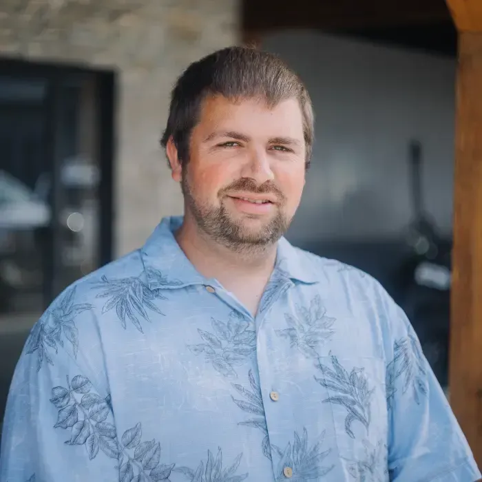 Man in a light blue Hawaiian shirt standing outdoors, smiling at the camera