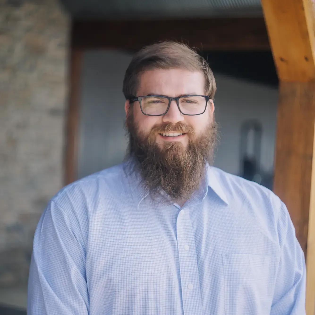 Smiling bearded man in glasses wearing a light blue shirt, standing outdoors by a wooden structure.