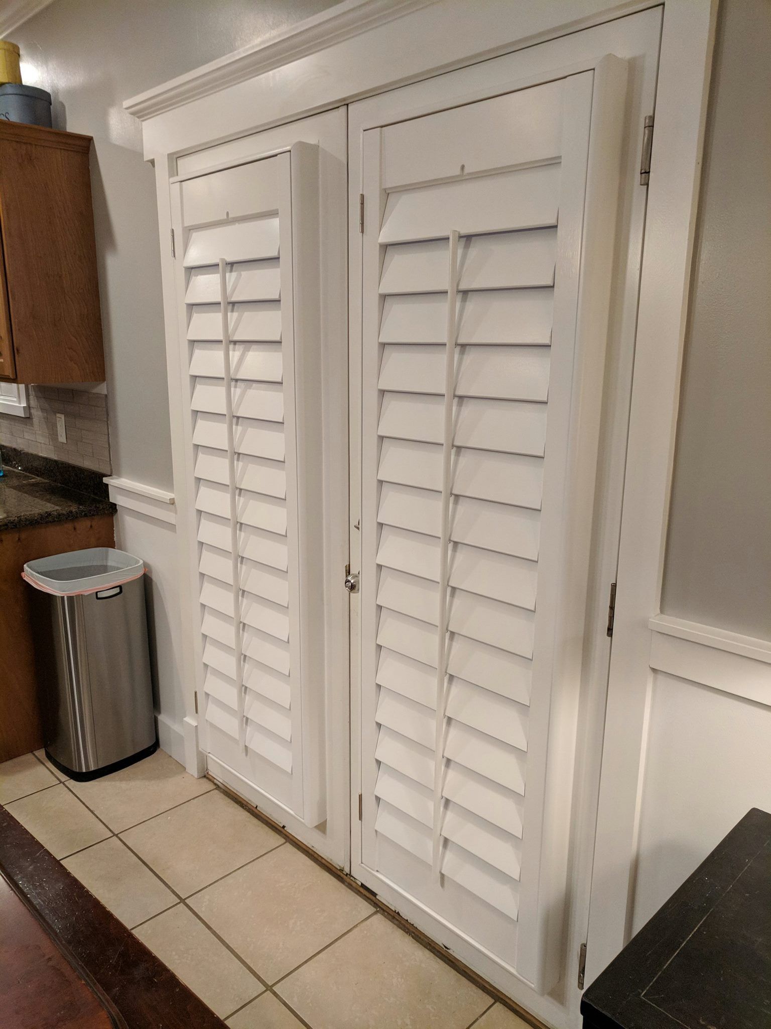 White louvered shutters on double doors within a white-trimmed doorway; kitchen setting with tile floor and a stainless steel trash can.
