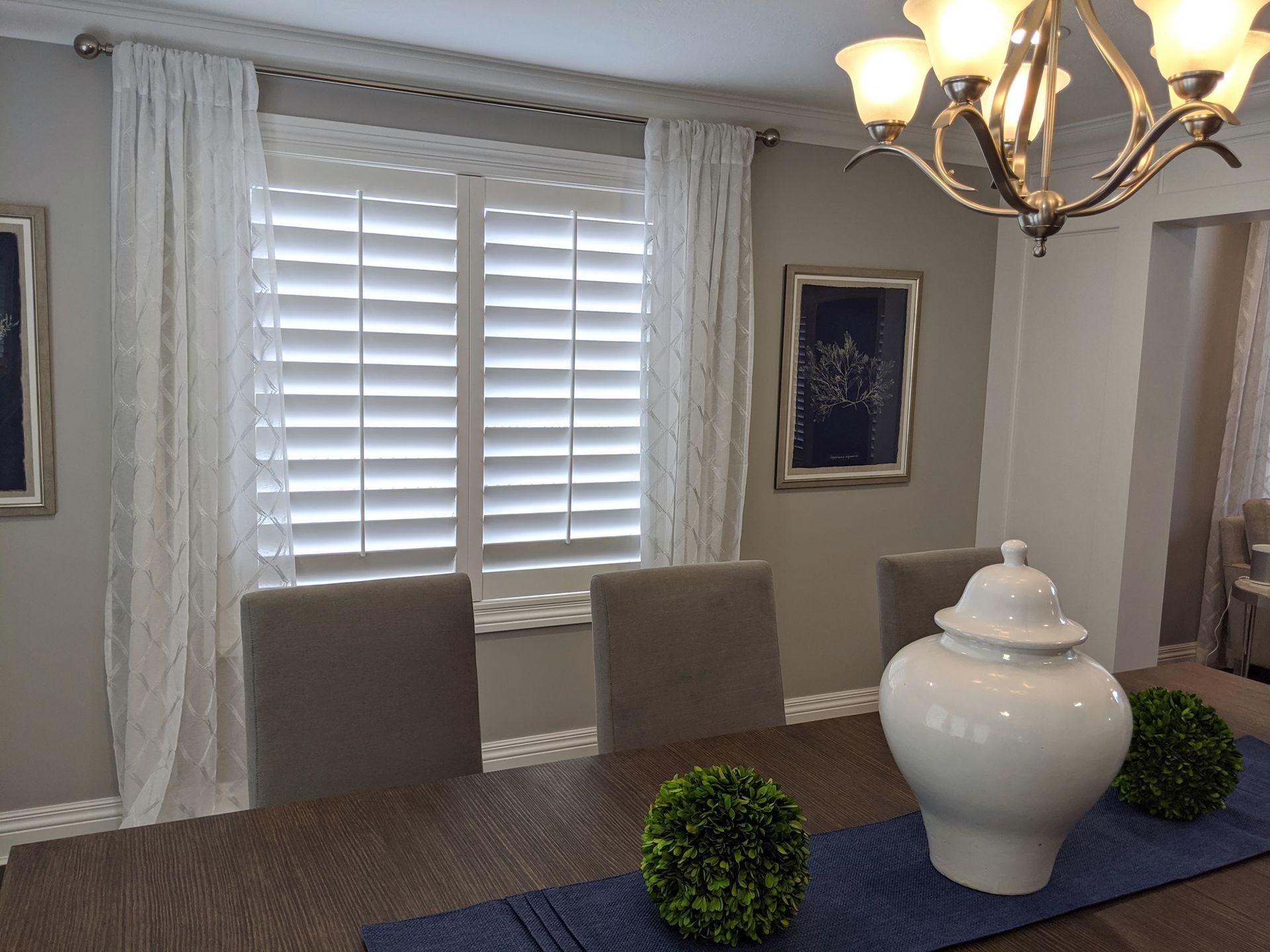 Dining room with table, white shutters, curtains, chandelier, and wall art.