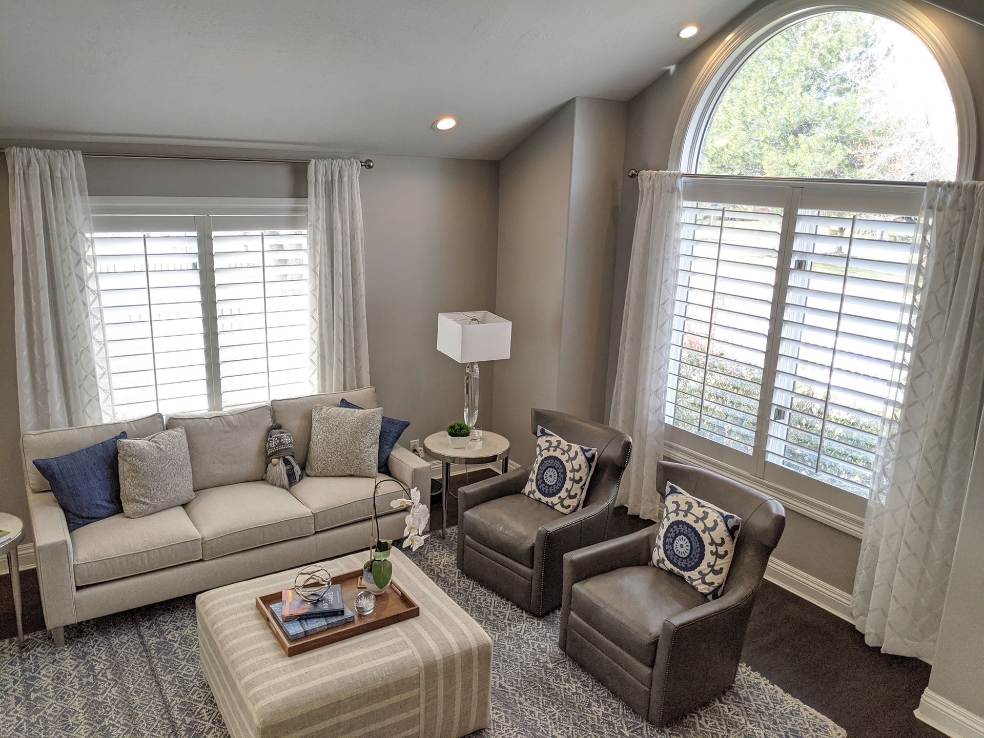 Living room with beige sofa, two leather armchairs, and large windows with shutters and white curtains.
