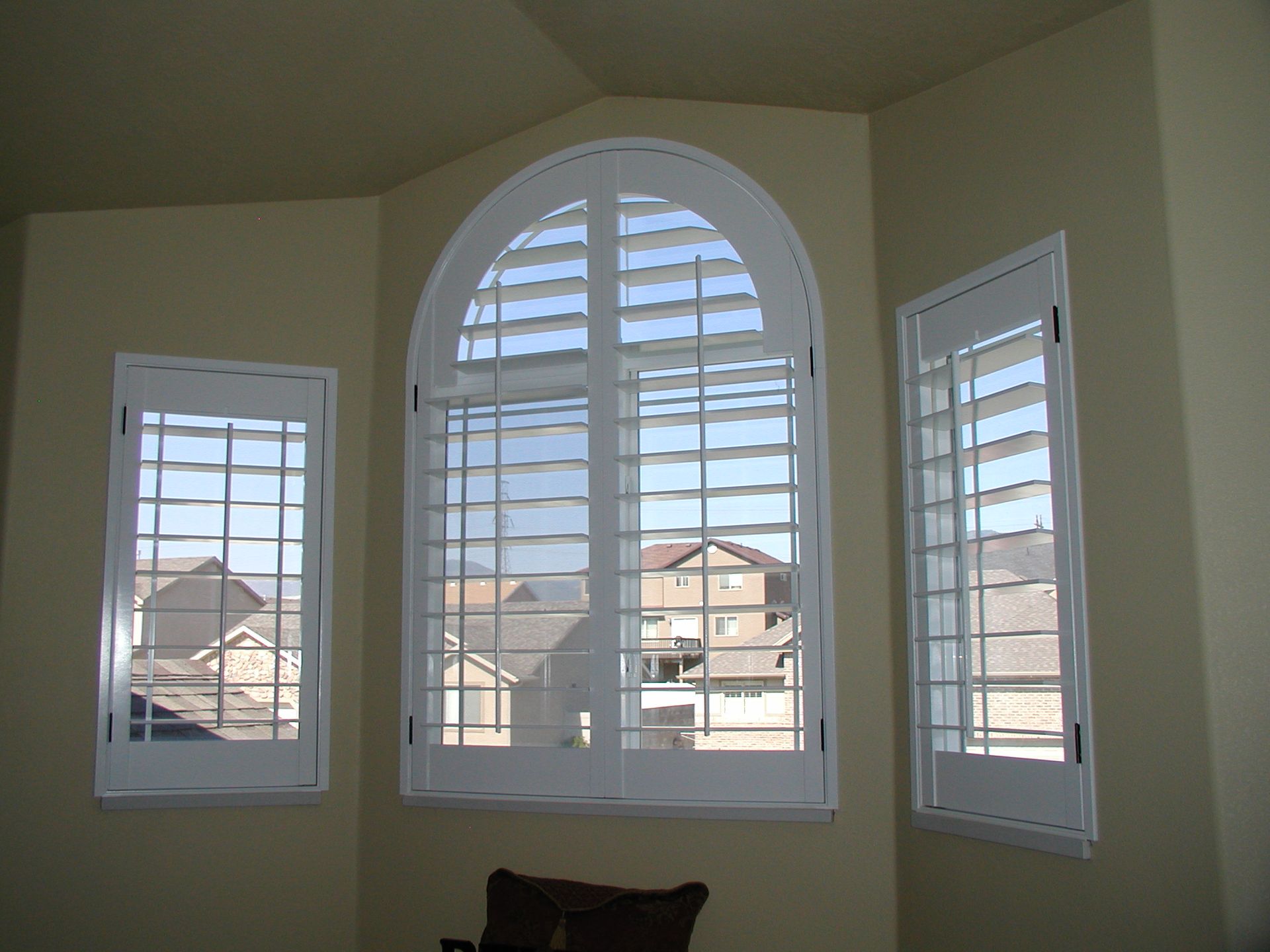 Three white-framed windows with white shutters in a tan-walled room, with a view of houses outside.