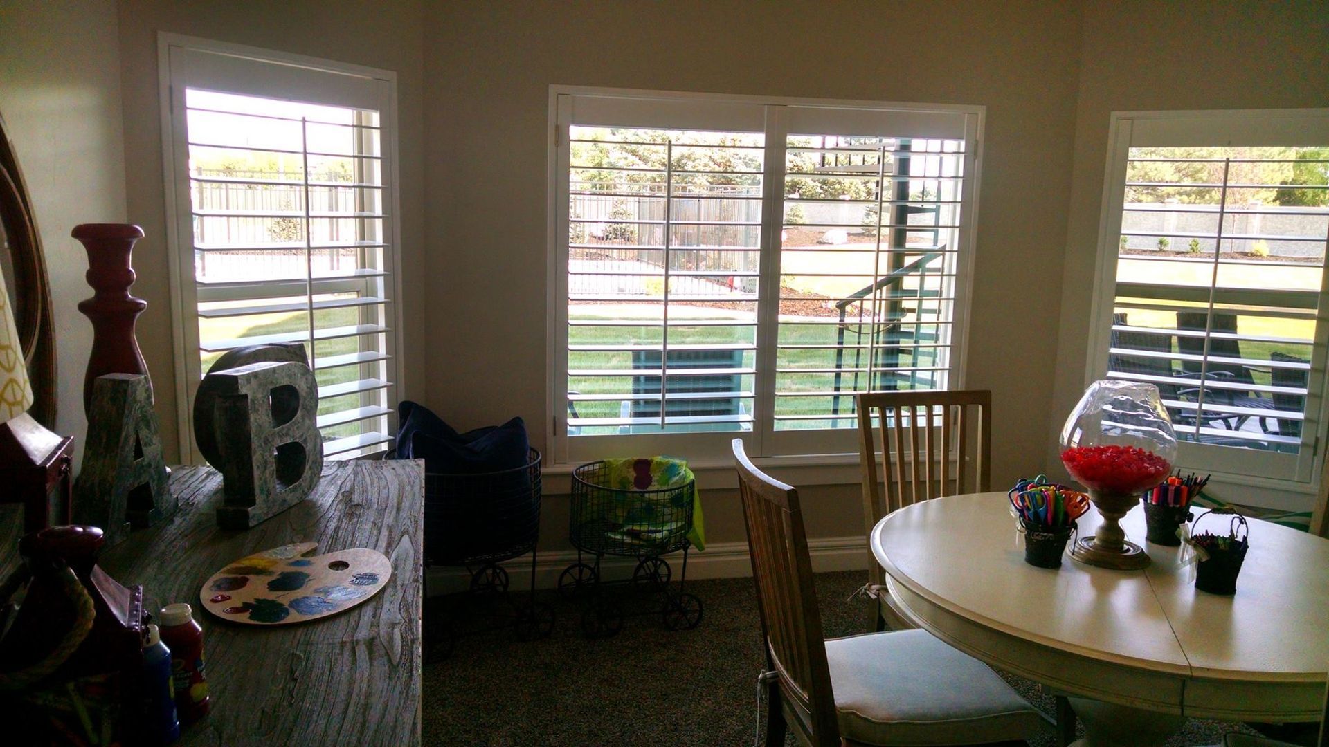 Room with shuttered windows overlooking a yard, table, chairs, and decorative items.