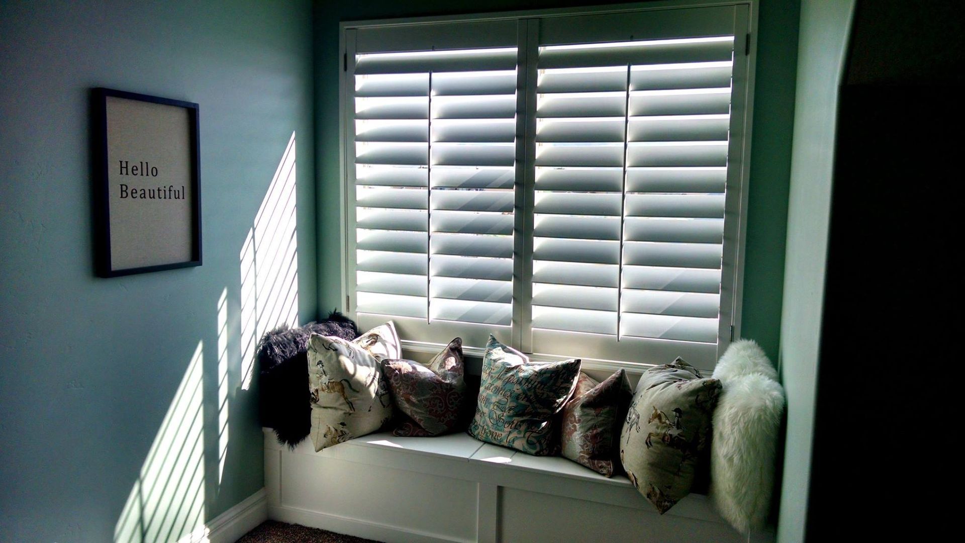 Window seat with white shutters and pillows against a blue wall. Black framed art on the wall.