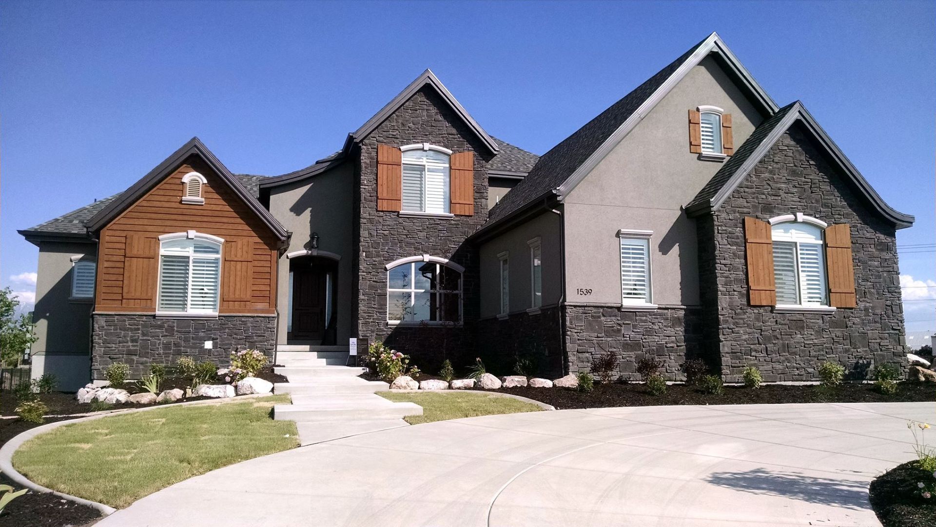 Two-story house with dark stone and brown shutters, gray stucco exterior, and landscaped yard on a sunny day.