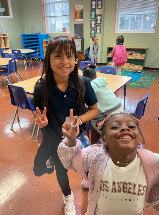 Two young girls are posing for a picture in a classroom. One girl is wearing a los angeles shirt.