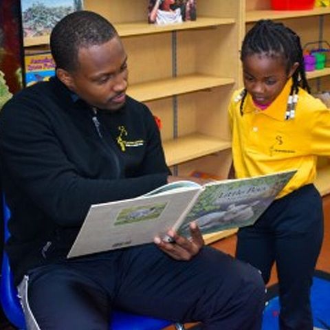 A man reads a book to a young girl in a classroom. Both are focused on the open book.