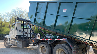 A dump truck with a green dumpster attached to it is parked in a gravel lot.