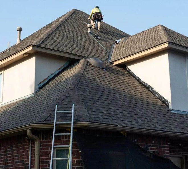 Roofer on a dark shingle roof, repairing it. Ladder against the house.