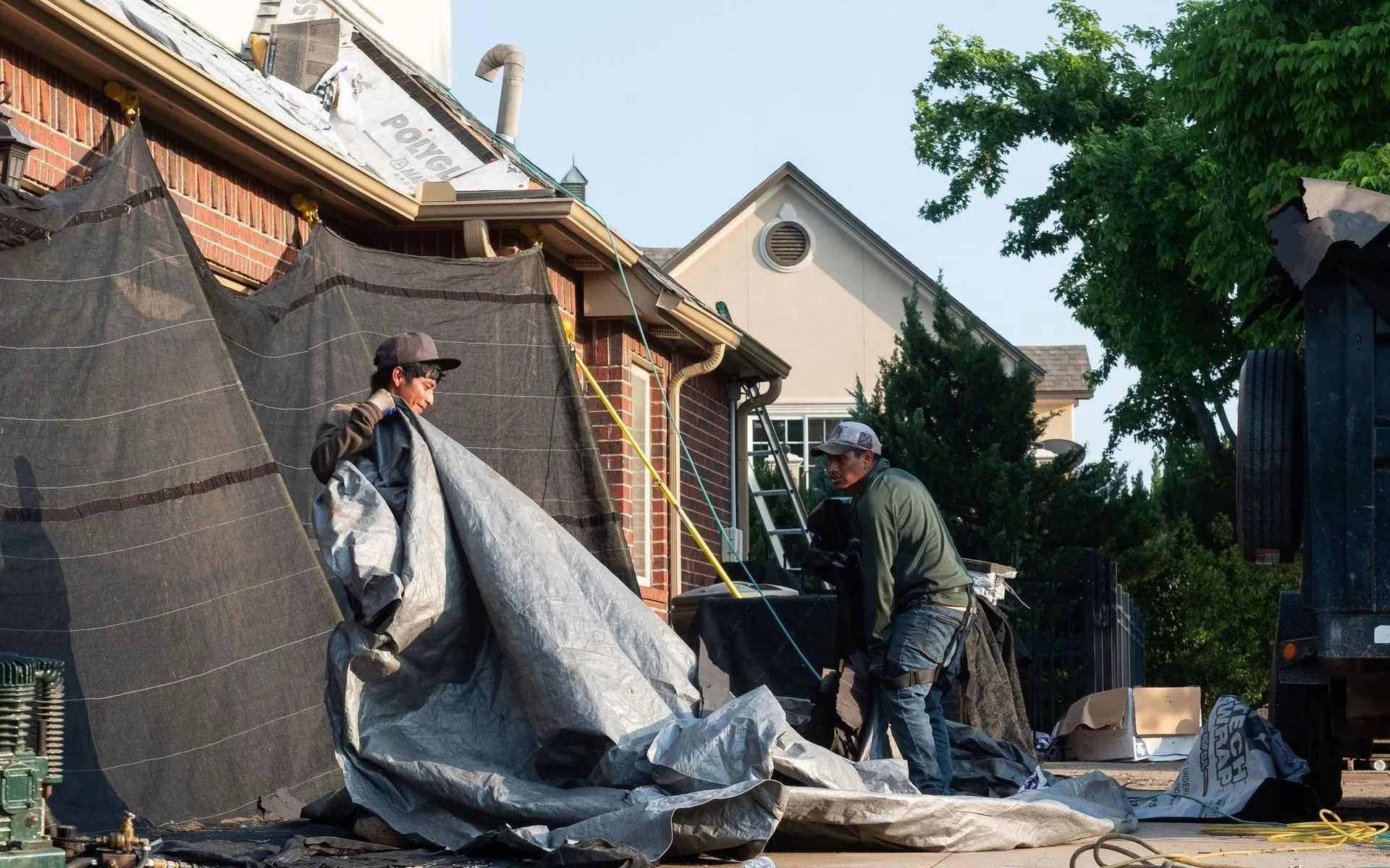 Two people are working on a building with tarps. A brick house and another building are in the background.