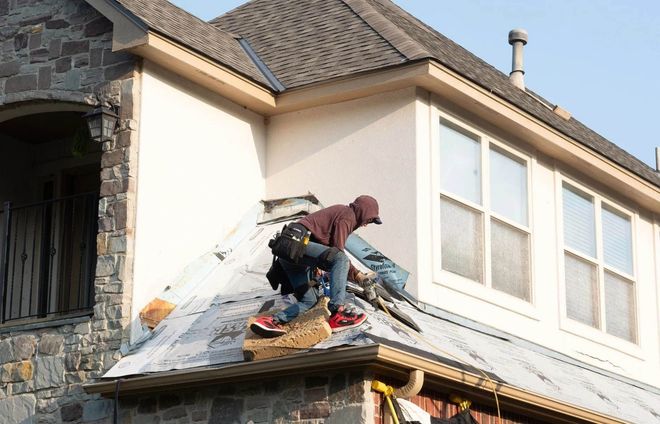 Roofer in hooded sweatshirt works on a roof, near windows and a stone wall.