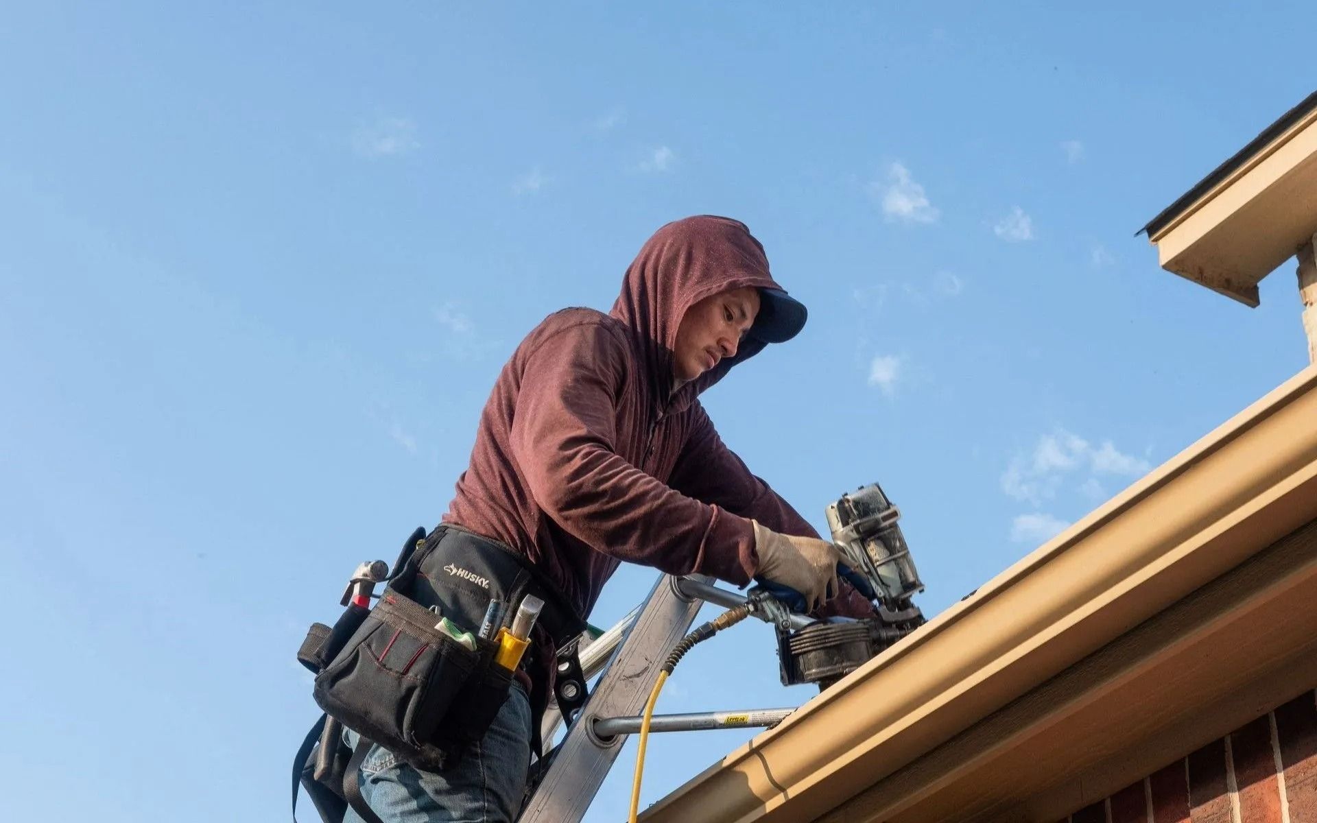 Person on ladder installing gutter, holding tool. Brown hooded sweatshirt, clear blue sky.