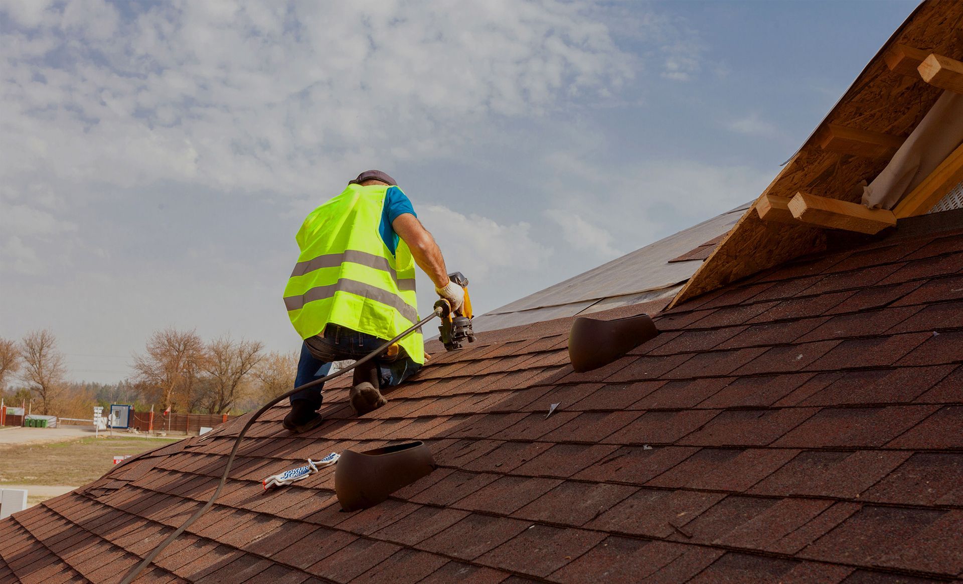 Roofer in yellow vest using a nail gun on a brown shingled roof under a cloudy sky.