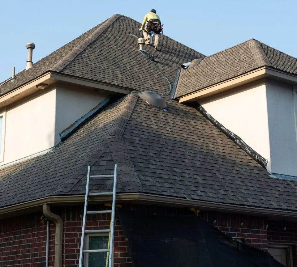 Roofer on a shingled roof, working near a chimney. Ladder leaning against the house.