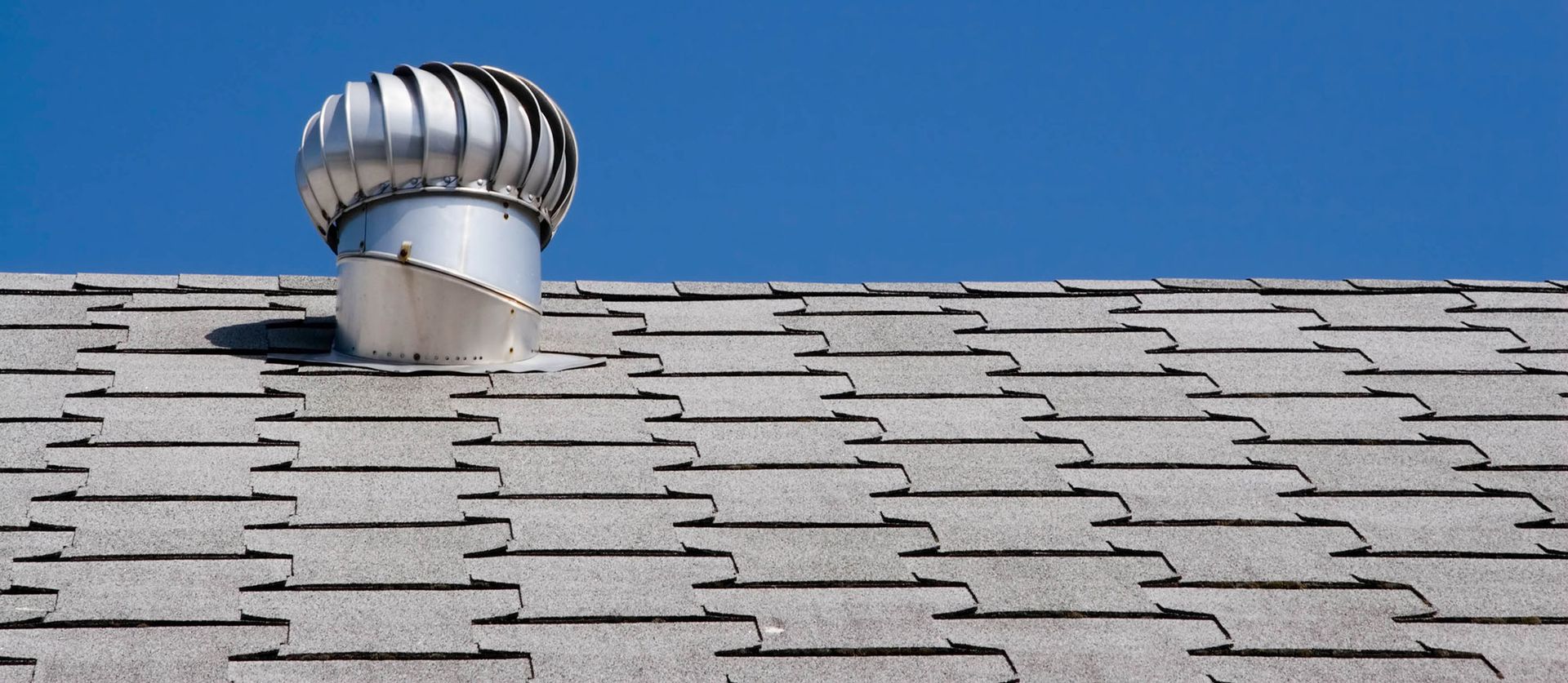 A silver roof turbine on a gray shingle roof against a blue sky.