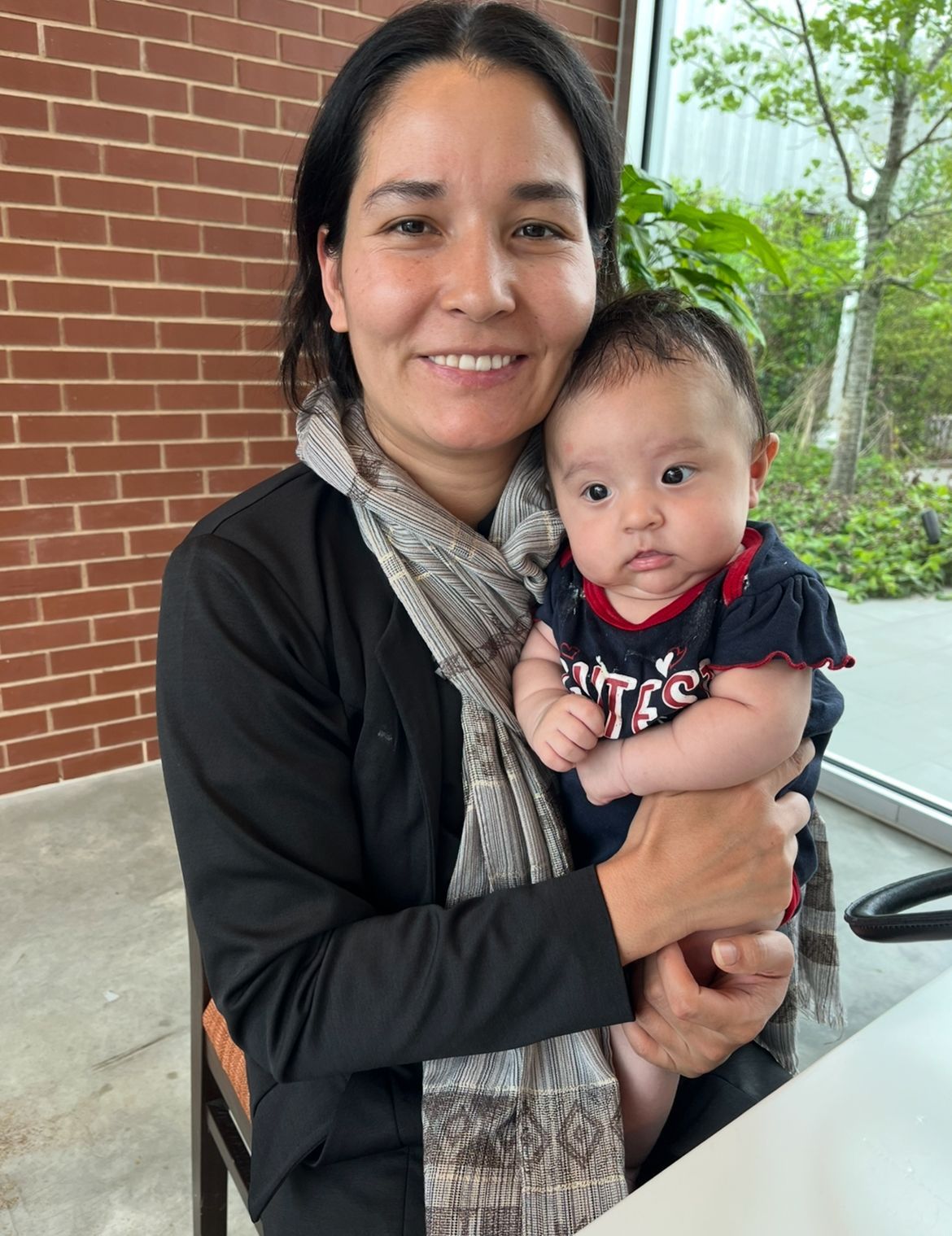 Woman holding a baby; both looking at the camera. Brick wall and greenery visible in the background.
