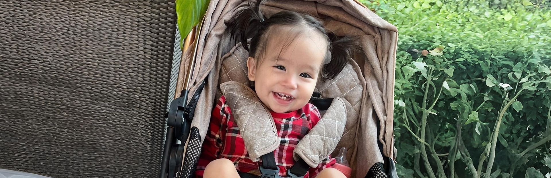 Smiling child in red plaid shirt and pigtails, secured in a stroller.