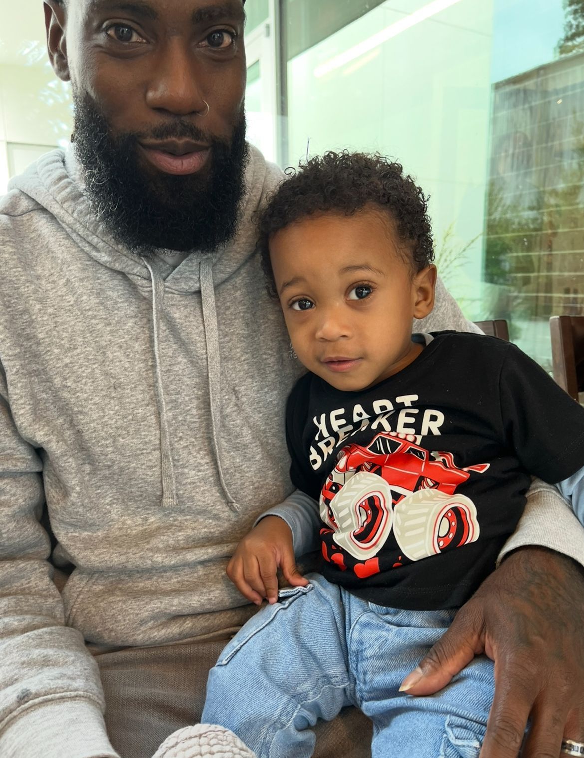 Man in gray hoodie holds a young child in a black shirt with a red design and blue jeans.