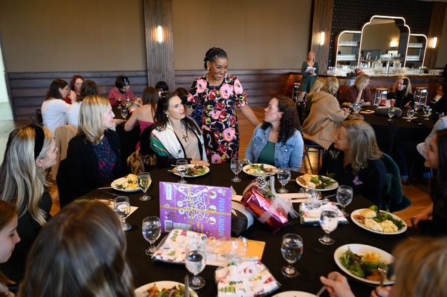 People at tables in a restaurant, a woman in floral dress standing. Black tablecloths, food, and glasses visible.