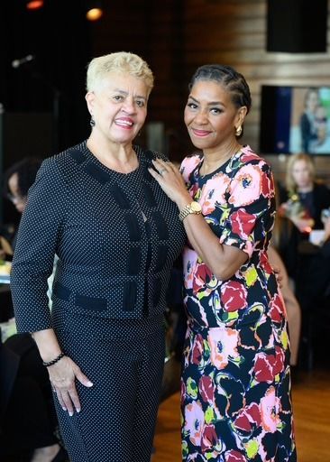Two women smiling, one in floral dress, the other in black suit. They stand inside, near a window.