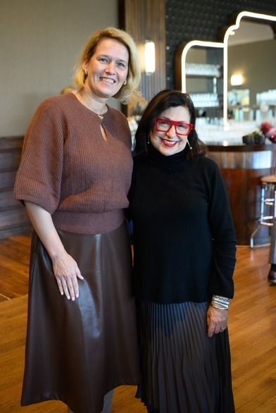 Two women smiling and posing indoors; one in brown sweater and skirt, the other in black turtleneck and red glasses.