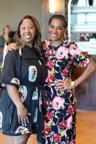 Two women smiling, posing for a photo. One in a floral dress, the other in a black dress, both indoors.