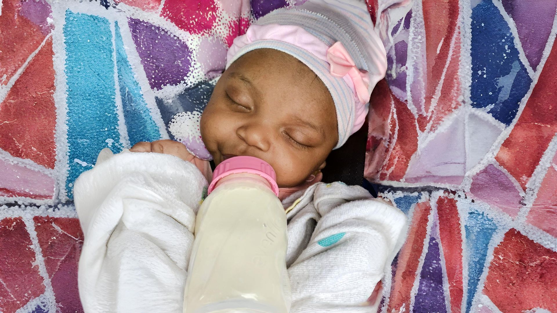 Baby sleeping while drinking from a bottle, wearing a pink bow hat, in a colorful blanket.