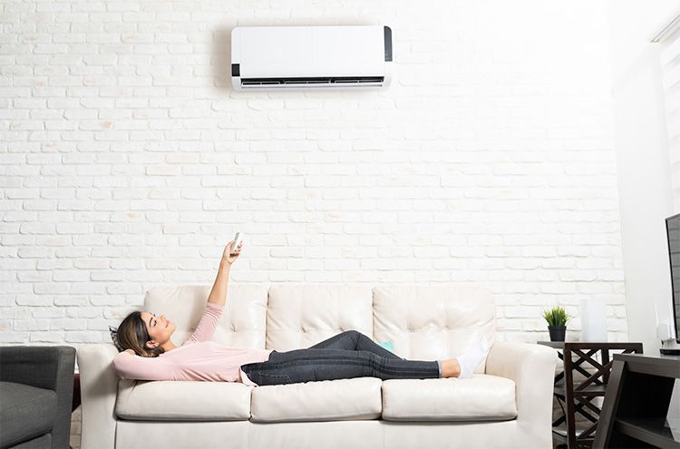 A person relaxes on a white couch while pointing a remote at a wall-mounted air conditioner in a brick-walled room.