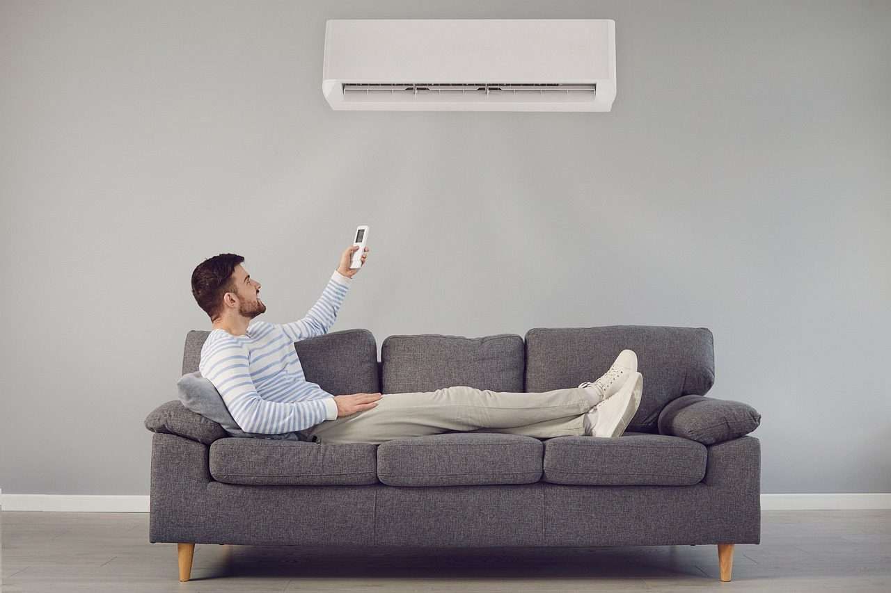 A person lounges on a grey couch, pointing a remote at a wall-mounted air conditioner.