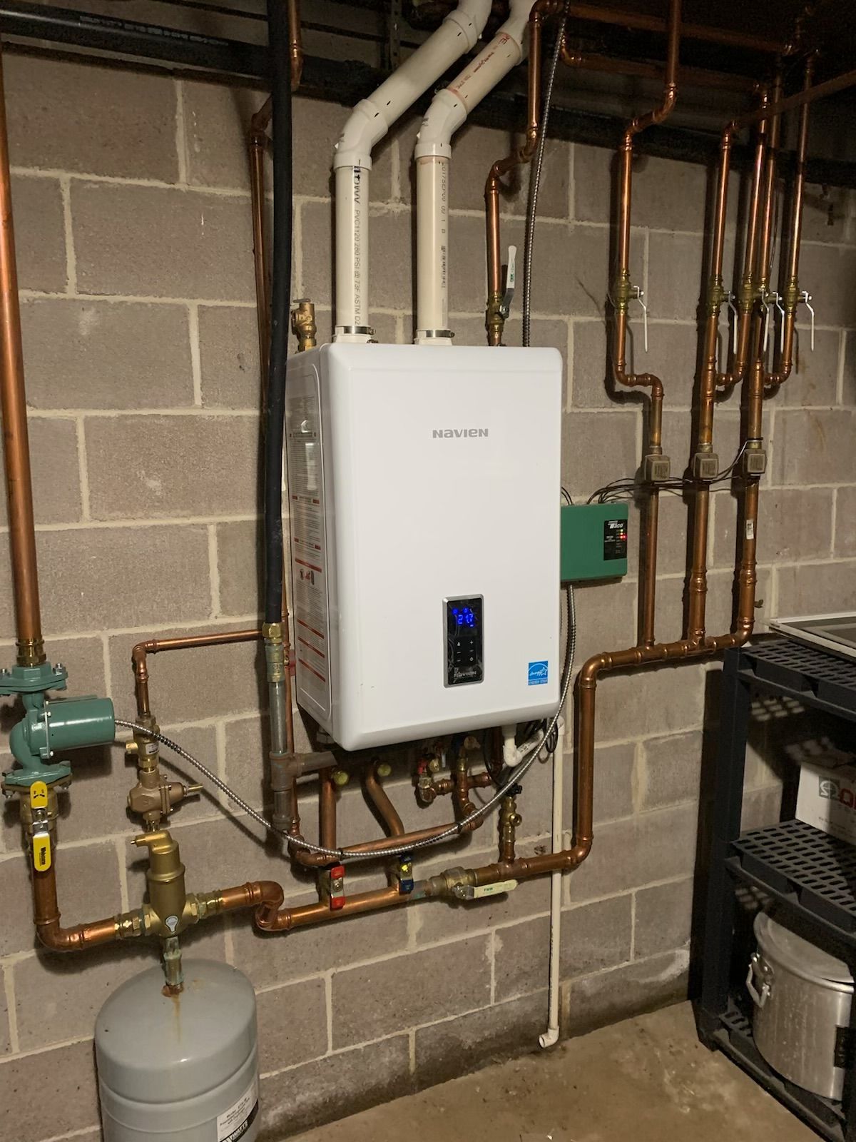 Wall-mounted white tankless water heater with PVC exhaust venting and connected copper piping in a basement setting.