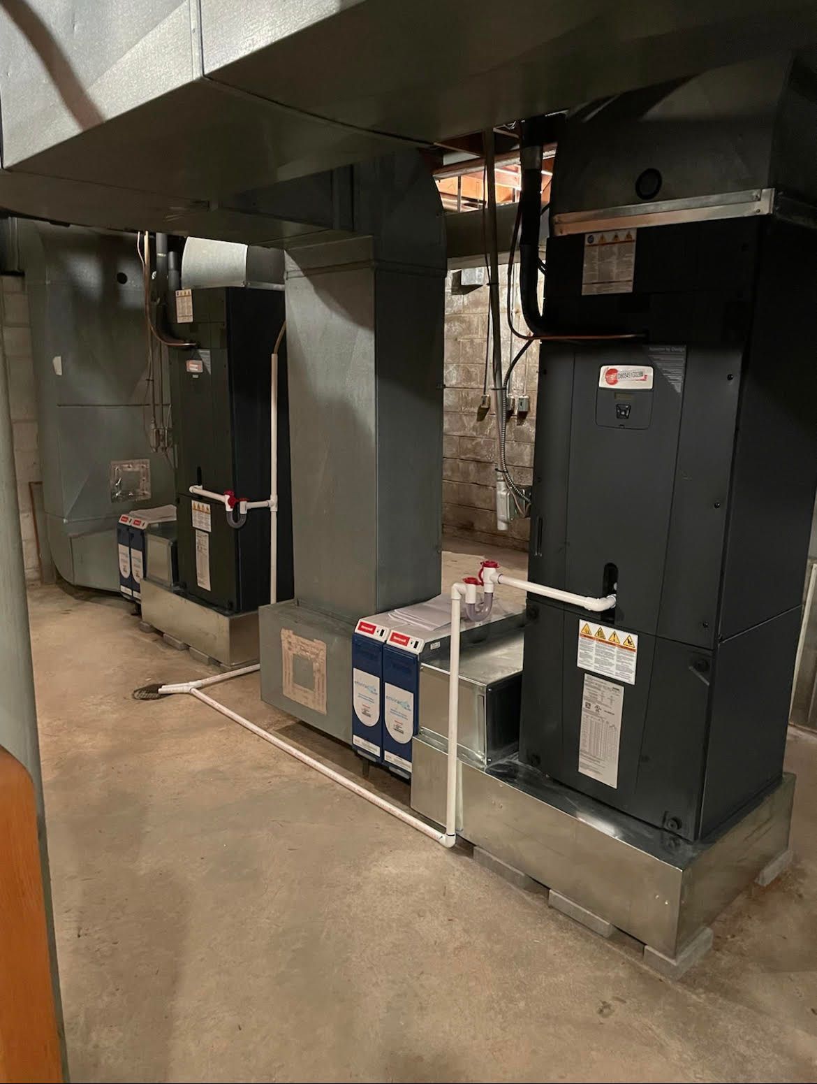 Two dark HVAC air handler units connected by metal ductwork and white PVC drainage pipes in a basement.