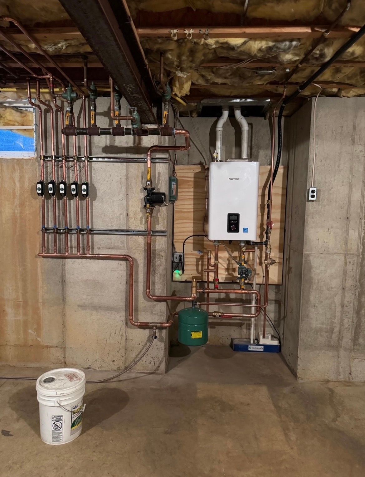 A wall-mounted tankless water heater with copper piping and a green expansion tank in an unfinished basement.