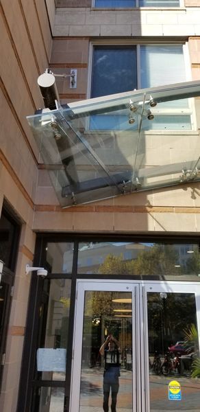 Glass awning over building entrance with reflective glass doors. Person taking photo in doorway.