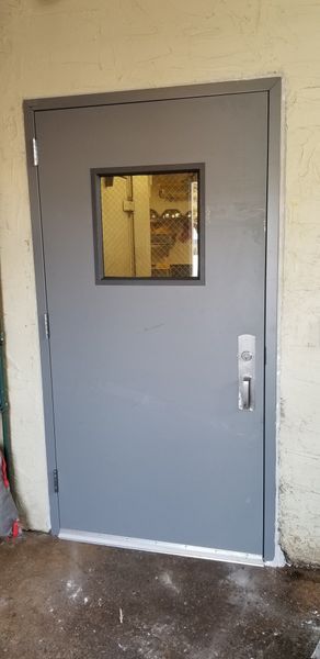 Gray door with a small window, handle, and lock, in an outdoor setting.