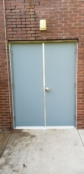 Two gray double doors in a brick building. Above the doors is a yellow light. Concrete ground below the doors.