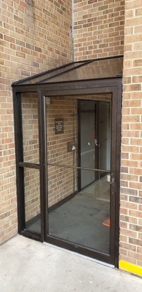 Glass-door entrance to a building with a brick wall. The door is open, and there is a small roof over the entrance.