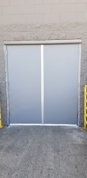 Closed gray warehouse doors. A white stripe runs vertically down the middle. Yellow barricades are visible on the right.