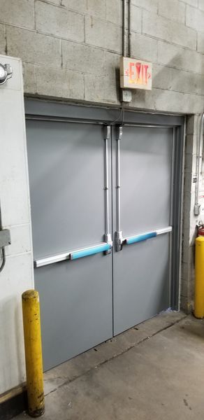 Gray double doors with exit sign above. Yellow bollards and concrete block wall.