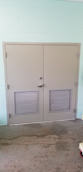 Gray double doors with vents in a concrete area against a light blue wall.