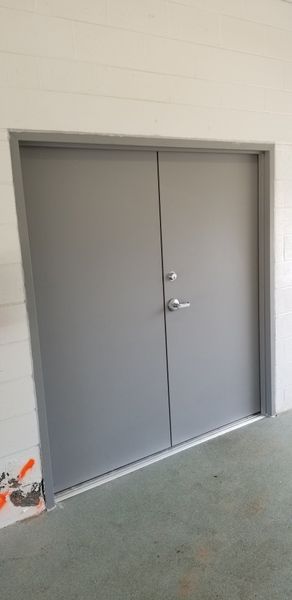 Gray double doors in a gray door frame, on a gray and green surface, against a white wall.