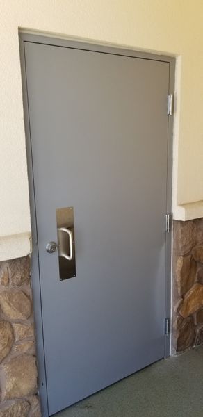 Gray metal door with a silver handle and doorknob in an outdoor setting with stone and light-colored walls.