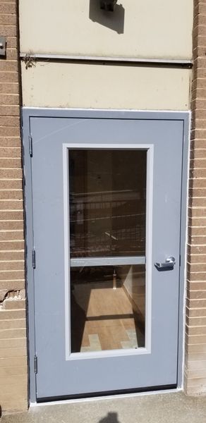 Gray door with window, set in brick building. Sunlight reflects on the glass, showing interior.