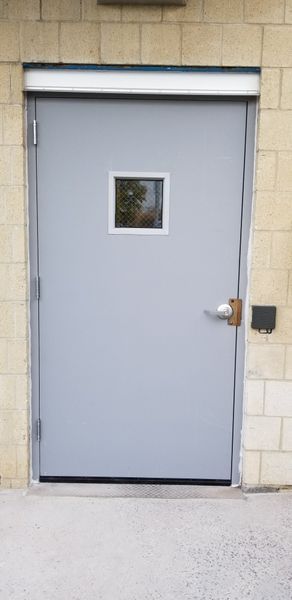 A gray metal door with a small square window, set in a light beige brick wall.