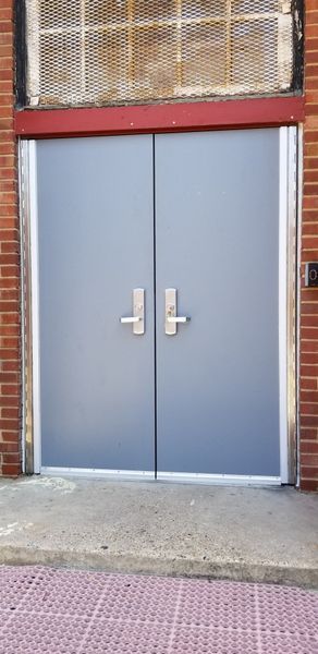 Two gray double doors with silver handles, set in a brick building. Above is a wire mesh window.