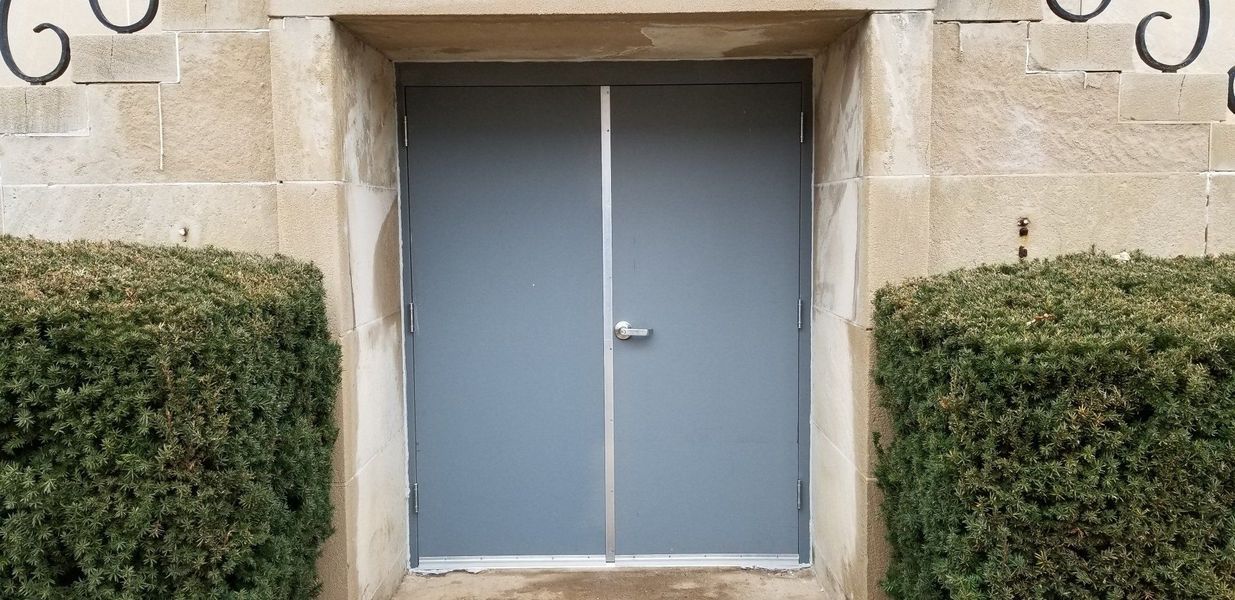 Gray double doors centered in a stone doorway, flanked by green bushes.