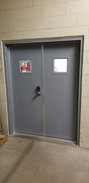 Gray double doors with small windows and a lock, set in a beige brick wall.